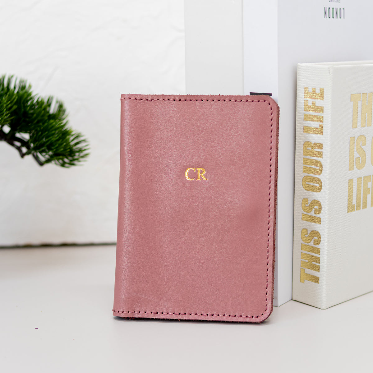 Pink leather passport wallet with gold embossed initials standing beside books
